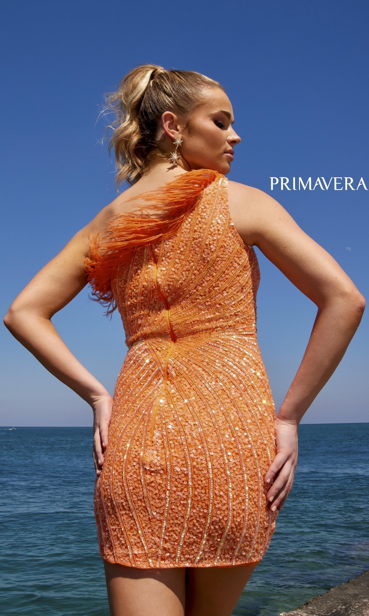 A woman stands by the ocean in the One-Shoulder Sequin Party Dress with Feathers 4002, featuring a vibrant orange hue, eye-catching sequins, and a feather-trimmed bodice for standout style.